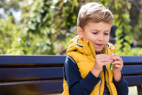 Parktaki bankta dinlenirken sakız paketini açmaya çalışan küçük bir çocuk..