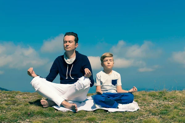 Zen benzeri bir baba ve oğul, çayırda meditasyon yapıyor ve gökyüzüne karşı dağ zirvesi yapıyor. Boşluğu kopyala.