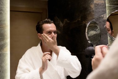 Banyoda yüzüne nemlendirici süren bir adamın aynadaki yansıması..