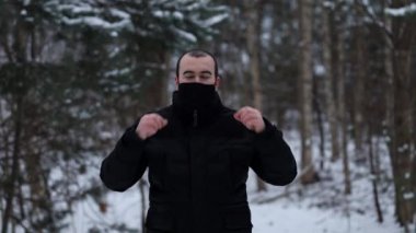 Dış giysili genç bir güneylinin portresinin görüntüsü, maskesini çıkarıyor ve tüttürüyor, kamera için sınıfını gösteriyor ve gülümsüyor, arka planda karla kaplı bir ormanda duruyor..