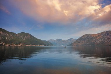 Karadağ 'ın Kotor Körfezi üzerinde sakin bir su, dağ yansımaları ve sakin bir kıyı manzarası yaratan sıcak bir sabah ışığı ile huzurlu bir gün doğumu.