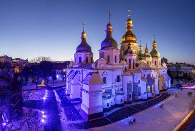 Kyiv 'deki Aziz Sophia Katedrali' nin kış akşamı manzarası gün batımından sonra, karların üzerinde parlayan altın kubbeler, derin mavi alacakaranlık gökyüzü ve yumuşak şehir aydınlığı altında..