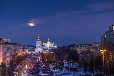Kyiv 'deki Sofya Meydanı' nda kış akşamı St. Michaels Altın Kubbeli Manastırı 'nın üzerinde yükselen dolunay, parlayan kubbeler, karlı sokaklar, şehrin ışıkları, sakin mavi gökyüzü, tarihi şehir atmosferi.