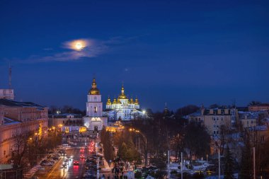 Kyiv 'deki Sofya Meydanı' nda kış akşamı St. Michaels Altın Kubbeli Manastırı 'nın üzerinde yükselen ay, parlayan kubbeler, karlı sokaklar, şehrin ışıkları, sakin mavi gökyüzü, tarihi şehir atmosferi.