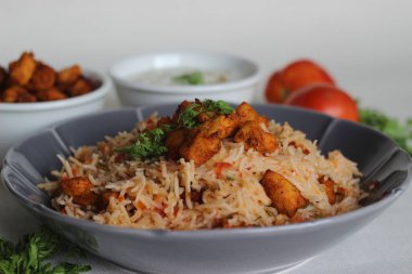 Raita ile servis edilen domates pilavı ve kızarmış kemiksiz tavuk ısırığı. Beyaz arka planda çekilmiş. Gri bir kasede servis edildi.