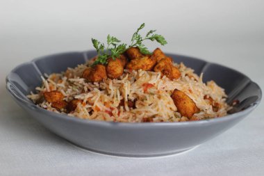 Raita ile servis edilen domates pilavı ve kızarmış kemiksiz tavuk ısırığı. Beyaz arka planda çekilmiş. Gri bir kasede servis edildi.