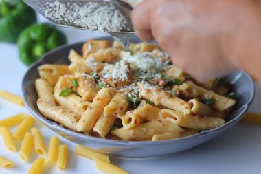 Hand grating cheese over creamy white sauce rigatoni pasta, with herbs, green bell peppers, and uncooked pasta in the background. Fresh Italian cooking concept with close up detail