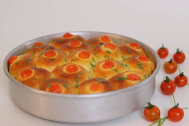 Çeri domates ve otlarla kaplı yuvarlak bir tavaya yerleştirilmiş taze pişmiş altın yemek ruloları, temiz beyaz bir arka planda çekilmiş ev yapımı ekmek ve konforlu yiyecek sergileniyor.