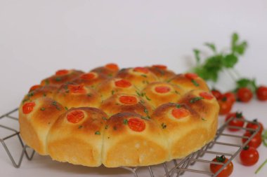 Çeri domates ve otlarla kaplı yuvarlak bir tavaya yerleştirilmiş taze pişmiş altın yemek ruloları, temiz beyaz bir arka planda çekilmiş ev yapımı ekmek ve konforlu yiyecek sergileniyor.