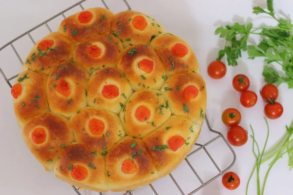 Çeri domates ve otlarla kaplı yuvarlak bir tavaya yerleştirilmiş taze pişmiş altın yemek ruloları, temiz beyaz bir arka planda çekilmiş ev yapımı ekmek ve konforlu yiyecek sergileniyor.