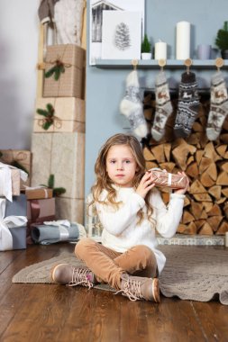 Beyaz kazaklı şirin bir kız şöminenin ve zarif bir Noel ağacının yanında oturur ve elinde kahverengi bir hediye, kameraya mutlu bir bakış atar ve ekolojik bir hediyeyi duymaya çalışır.