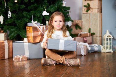 Beyaz kazaklı ve kahverengi pantolonlu şirin bir kız şöminenin yanında oturuyor ve Noel ağacını giydirip elinde büyük gri bir hediyeyle kameraya bakıyor.