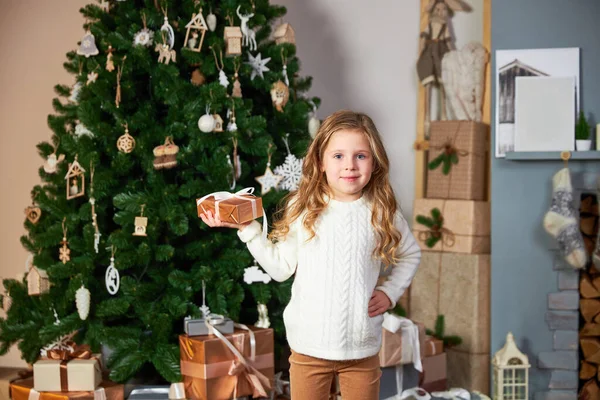 Słodka dziewczynka w białym swetrze stoi obok eleganckiej choinki i posiada zielony prezent ekologiczny, patrzy w aparat i uśmiecha się — Zdjęcie stockowe