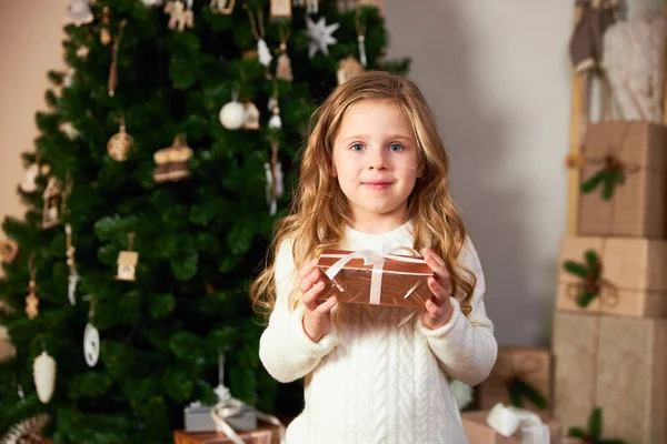 Słodka dziewczynka w białym swetrze stoi obok eleganckiej choinki i posiada zielony prezent ekologiczny, patrzy w aparat i uśmiecha się — Zdjęcie stockowe
