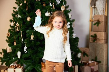 Beyaz kazaklı ve eko elbiseli bir kız Noel ağacı ve hediyelerin yanında duruyor. Kızın elinde tıbbi bir maske var, maskesini çıkartıyor. Kız gülümsüyor.