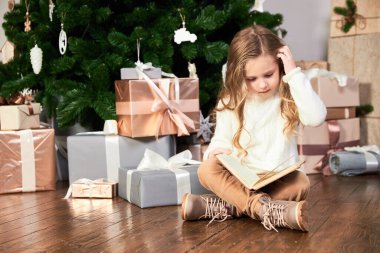 Beyaz elbiseli bir kız Noel ağacının yanına oturur ve bir kitap okur, kafasını kaşır ve düşünür.
