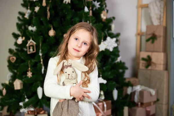 A little cute girl stands against the background of a green Christmas christmas tree and holds a toy hare — Stock Photo, Image