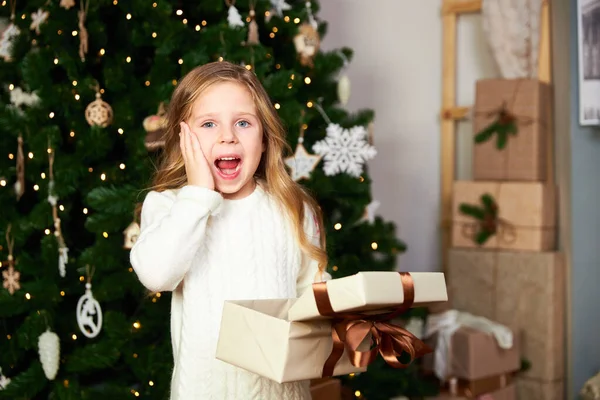 Het meisje staat in een kamer met een groene kerstboom en houdt een geschenk, het meisje kreeg een cadeau voor Kerstmis en gelukkig, het meisje kijkt in de camera — Stockfoto