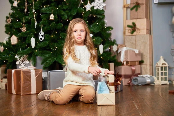Dziewczyna wyciąga maskę medyczną z prezentu świątecznego. Dziewczyna jest zdenerwowana i patrzy w kamerę. — Zdjęcie stockowe