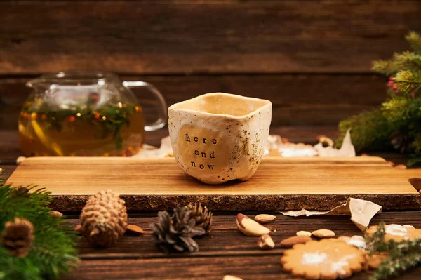 Dezember, Weihnachten. Holzhintergrund, Plätzchen, Kegel und Nüsse. Ein Keramikbecher mit der Motivation im Hier und Jetzt. — Stockfoto