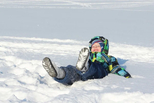 Маленький хлопчик в кольоровій куртці з капюшоном лежить на снігу — стокове фото