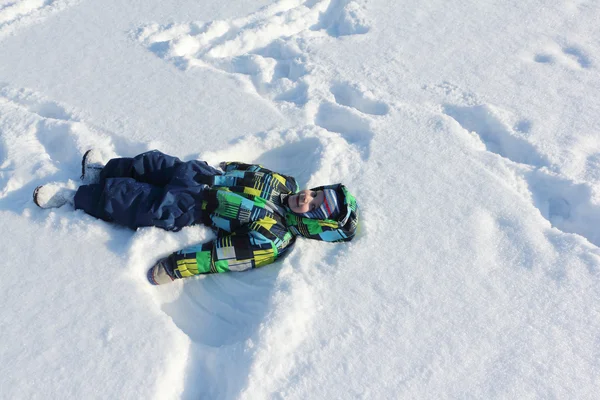Il bambino in una giacca a colori con un sorriso sdraiato sulla neve — Foto Stock