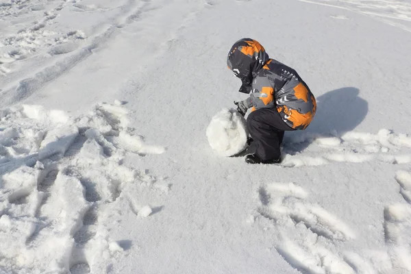Il bambino in una giacca a colori con un cappuccio che fa rotolare una palla di neve — Foto Stock