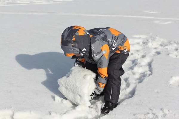 El niño en una chaqueta de color con una capucha rodando una bola de nieve —  Fotos de Stock