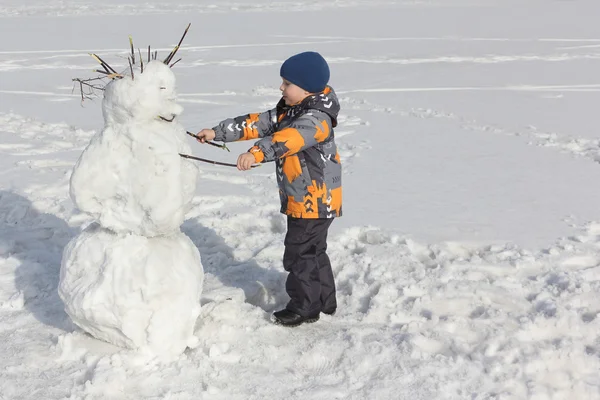 O menino em uma jaqueta colorida construindo um boneco de neve — Fotografia de Stock
