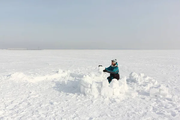 Happy woman in a green jacket building an igloo on the snow in winter, Ob reservoir, Novosibirsk, Russia