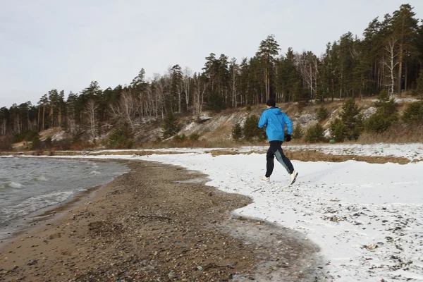Ο άνθρωπος σε ένα μπλε σακάκι που τρέχει στο χιόνι στην όχθη του ποταμού — Φωτογραφία Αρχείου