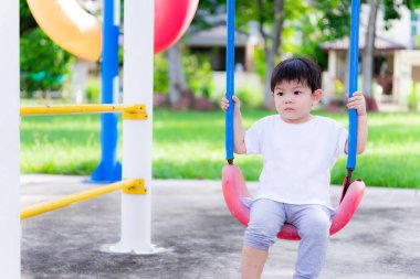 Asyalı anaokulu çocuğu kırmızı salıncaklarda oturuyor, yalnız ve üzgün hareketler yapıyor. Korkmuş küçük bir çocuk 2 yaşında. Çocuk parkında oynayan bebeğin yeşil bir arka planı var..