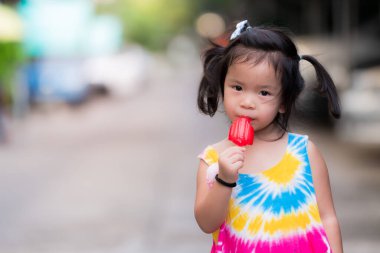 Kız yürüyüşe çıkar ve kırmızı çubuklu dondurma yer. Çocuk renkli kolsuz gömlek giyiyor. Asyalı çocuk kameraya bakıyor. İyi huylu bir çocuk, hafif tatlı bir gülümseme. 3 yıl 5 aylık çocuklar.