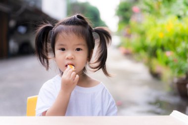 Sevimli kız portakal yiyor. Çocukların meyve yemesi zordur. Çocuk beyaz gömlek giyiyor. Öndeki küçük tahta masa. İki siyah saçı olan sevimli bir çocuk. Yeni yürümeye başlayan çocuk 3 yaşında..