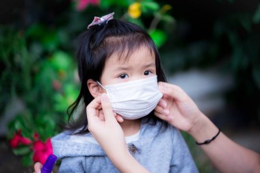 Beyaz tıbbi maske takan bir çocuk onun üstündeki bir şeye bakıyor. Ailesinin elleri maskeleri, Asyalı kızların virüs ve tozla rahat edebilmeleri için diziyor..