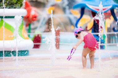 Çocuk çeşmeye bakıyor. O, kameraya sırtını döndü. Asyalı kız kırmızı mayo giyiyor ve sol elinde mor yüzme gözlüğü tutuyor. Güneşli bir günde oynayan bir çocuk. 3 yaşında çocuklar..