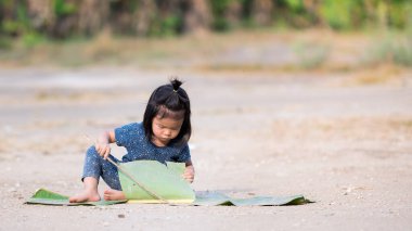 Çocuk yerde oturup toz toprak dallarla ve muz yapraklarıyla oynuyor. Çocuklar doğayla oynar. Sıcak bir günde sıcaktan terleyen bir çocuk. Mavi elbiseli küçük kız 2 yaşında..