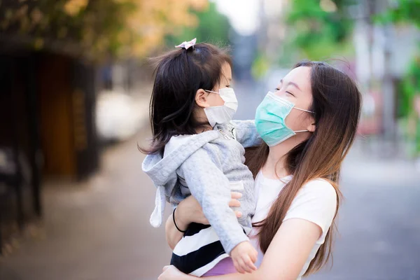 Concept of mother caring for daughter wearing surgical medical face mask. Mother carried and embraced daughter with love on public road. Both of them wear masks to protect against dust and viruses.