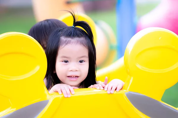 遊び場で かくれんぼ をしているアジア系の女の子 就学前の子供は友人や彼女の兄弟と黄色のプレーヤーマシンの後ろを覗きます 幸せな子供たち遊びで甘い笑顔 — ストック写真