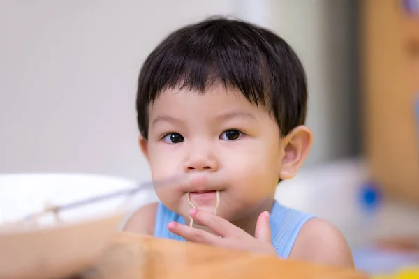 2歳の少年は インスタントラーメンを拾うために手を使います アジアの赤ん坊の男の子は彼の口の中にインスタントラーメンの行を入れて カメラを見た 息子は家で食べ物を食べている — ストック写真