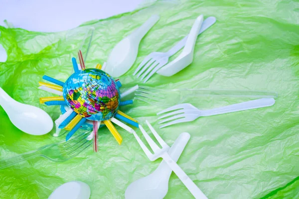 World is full of many plastic waste. Water tube is plugged in the middle of the globe. World ball is placed on the green bag. White and clear plastic spoons and forks are placed around the earth.