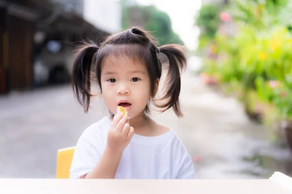 眠い目の子供が座ってオレンジを食べている 愛らしい少女は花弁を口に入れた かわいいです3歳アジアの子供を身に着けています白いドレスと2つの髪のネクタイ — ストック写真