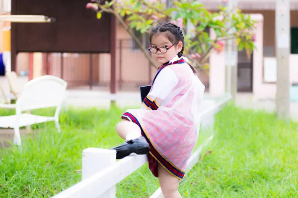 学校の制服を着たかわいいアジアの小さな子供の女の子は 遊び場で遊ぶために横に白い低いフェンスを登ることです 卒業式のドレス 6歳のお子様は眼鏡をかけて — ストック写真