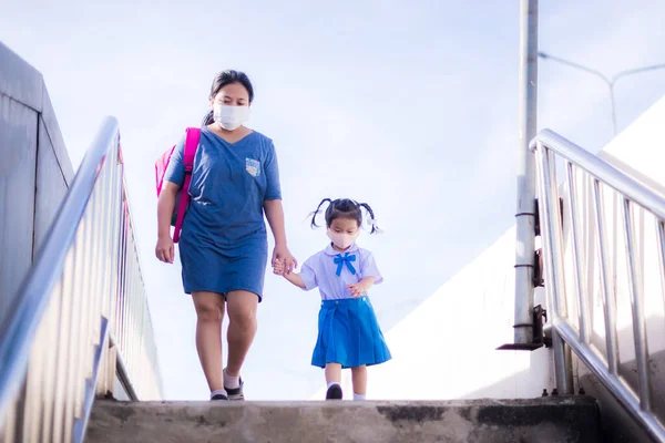 Mother and daughter wear cloth masks. Two of them walked hand in hand, walking down the steps of overpass to go to school. Mom carries pink school bag to help Asian girl. New normal concept of life.