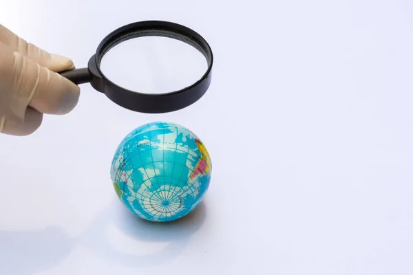 Person holding magnifying glass shines down onto the globe that is a ball made of foam or plastic. Person wearing the white gloves. Isolated white background. Concept of pollutant survey on our planet