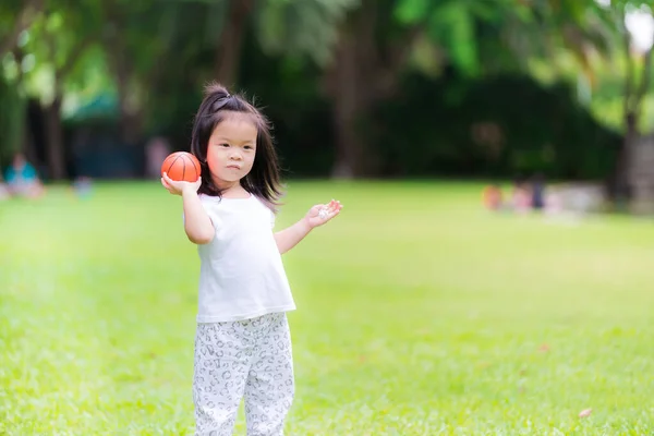 公園の緑の芝生で遊んでいる愛らしいアジアの少女は 小さなバスケットボールに似たオレンジ色のボールを投げます 幸せな子供 甘い笑顔 白い服を着て — ストック写真