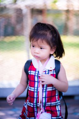 Dikey görüntü. 2-3 yaşındaki bir bebeğin portresi. Asyalı kız durdu ve kaşlarını çattı. O, içme suyu şişesinin kayışını kavradı. Ekose desenli kırmızı bir elbise giyiyor. Bir şeye bakıyor..