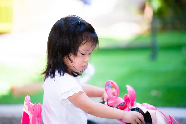 子供たちはピンクの電動おもちゃバイクを再生します みすぼらしい顔の子供 子供は怪しい表情をした 白いシャツを着た3歳の女の子が家の前で遊んでいる — ストック写真