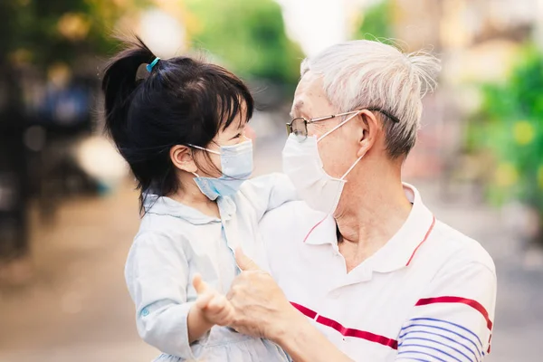 Family wear surgical face masks to prevent the spread of coronavirus. Grandfather held granddaughter talked, smiled and laughed. Adult and child look at each other and smile sweetly under medical mask