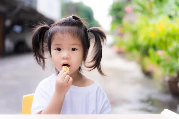 女の子は果物を食べることを余儀なくされた かわいい子供が家の前でオレンジを食べている 親は風邪を防ぐためにビタミンCを加えるためにオレンジを食べることを強いられる 悲しい顔の子供3歳の白いシャツを着て — ストック写真